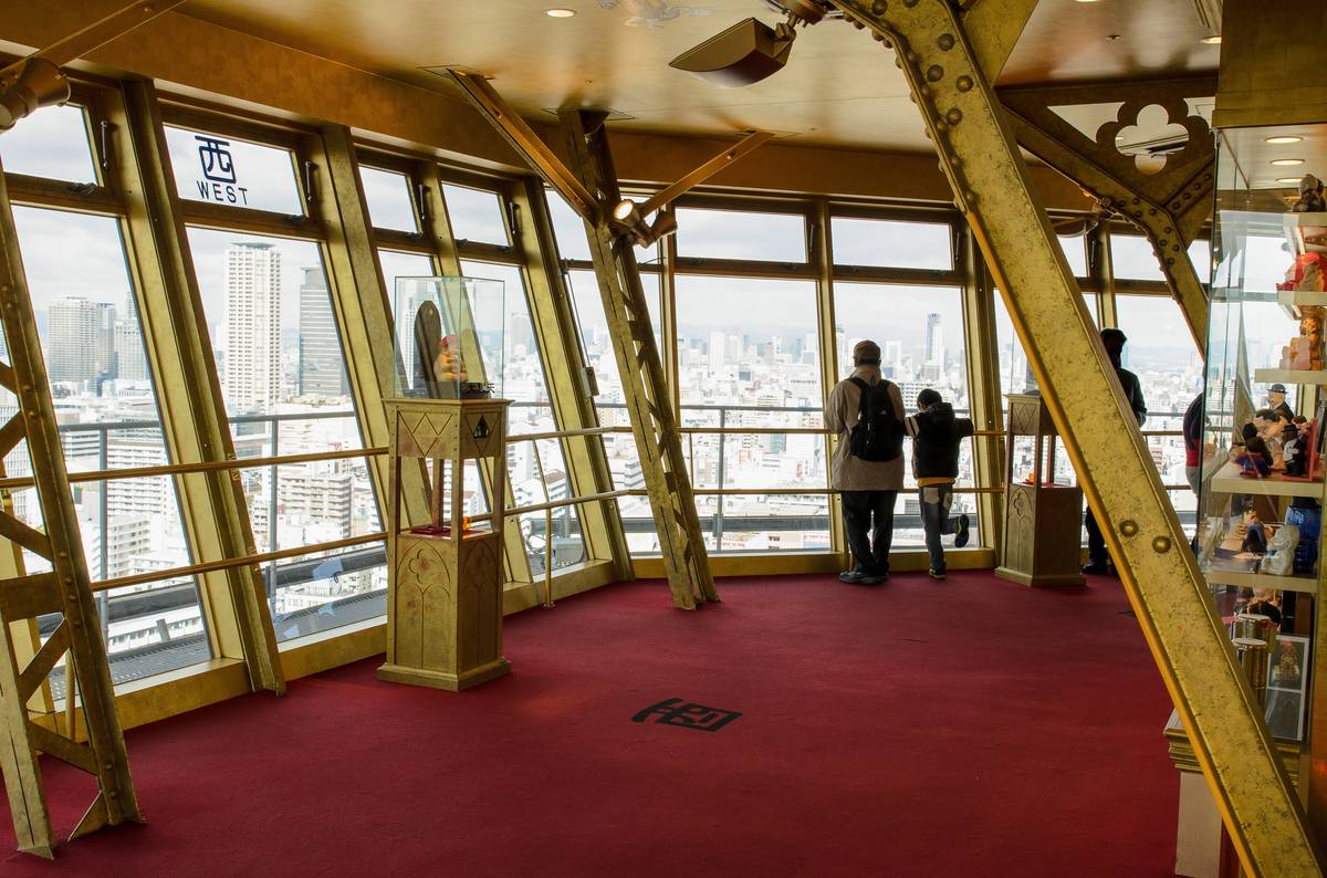 Observation deck with a red carpet, gold metal beams, a display case, and people looking out at a city view.