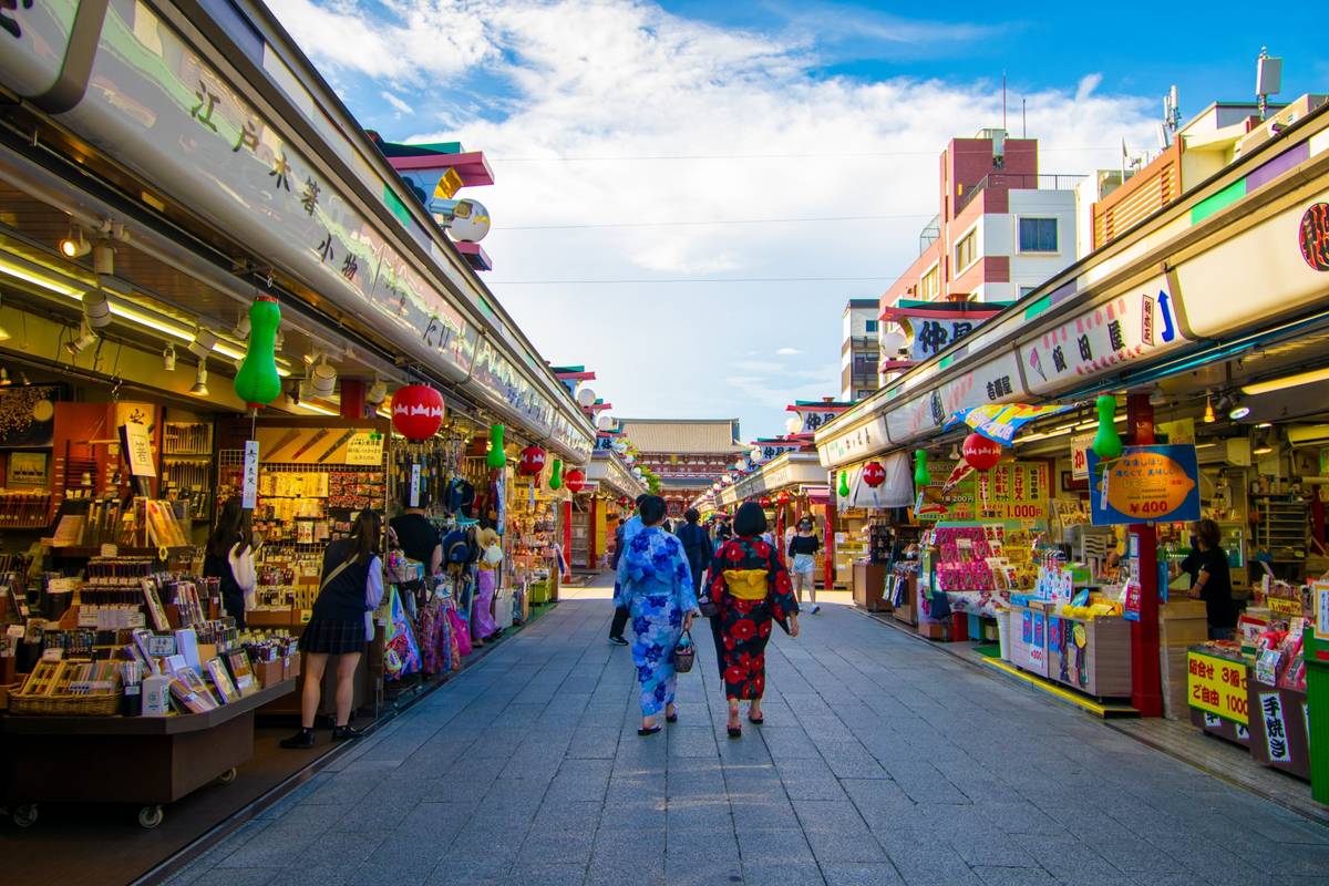 A lively outdoor market street in Japan, with rows of shops and people, some in kimonos, walking.