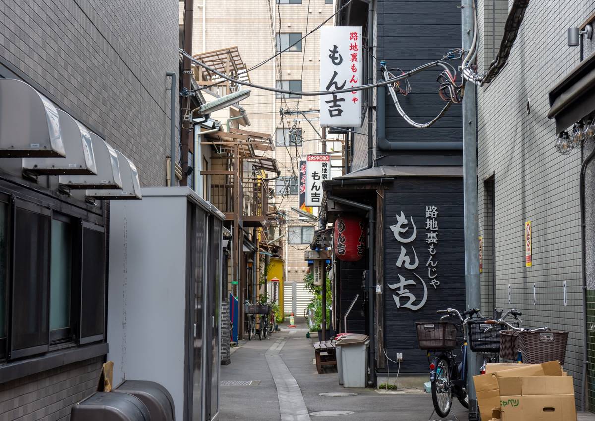 A narrow urban alley in Japan, lined with buildings, overhead utility wires, and a black storefront with white Japanese script, and parked bicycles on the right.