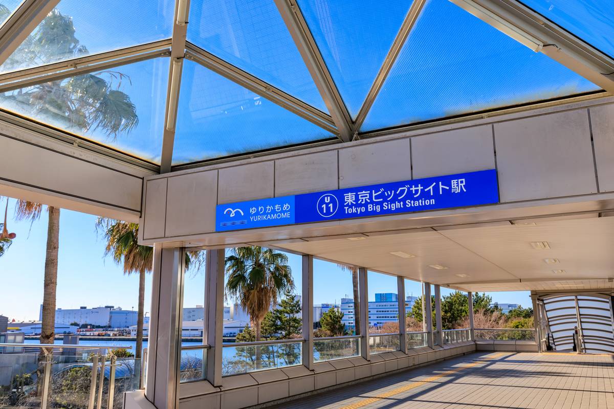 Elevated glass walkway with a Tokyo Big Sight Station sign, palm trees, and bay.