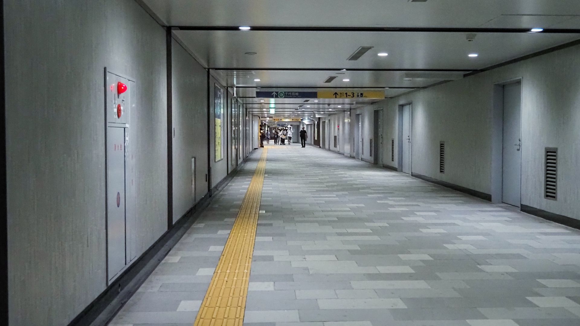 A long, modern underground hallway with grey tiled floor, a yellow tactile path, and multiple doors along the right wall.