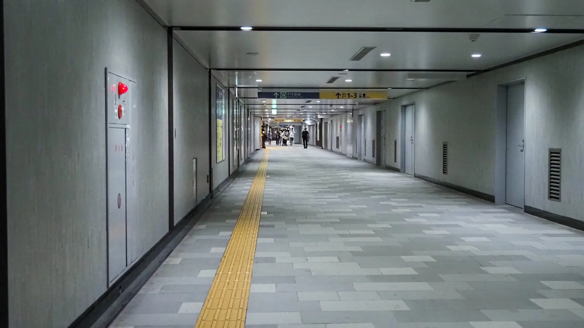 A long, modern underground hallway with grey tiled floor, a yellow tactile path, and multiple doors along the right wall.