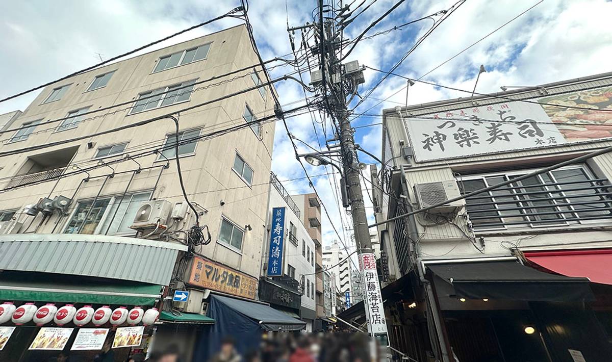 A bustling Japanese street lined with buildings and shops, crisscrossed by tangled overhead wires.