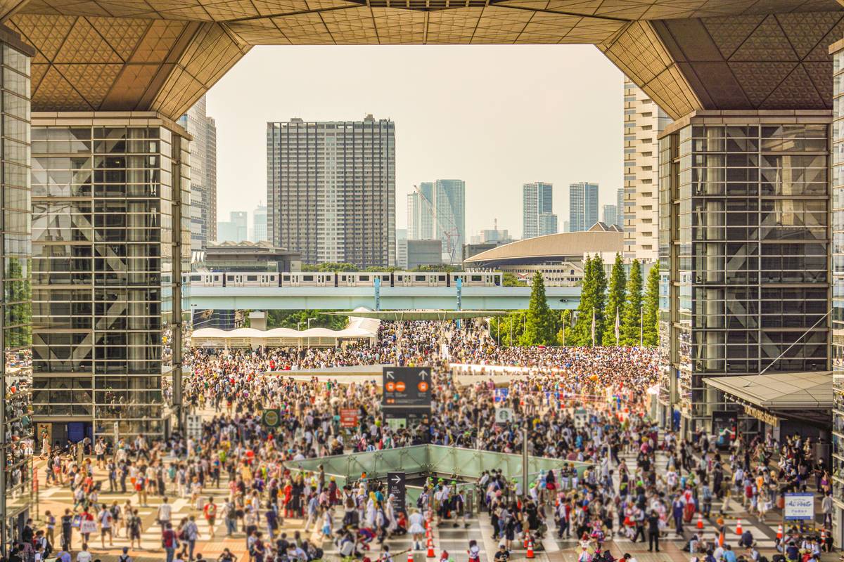 Massive crowd of people in an urban plaza with modern buildings and an elevated train.