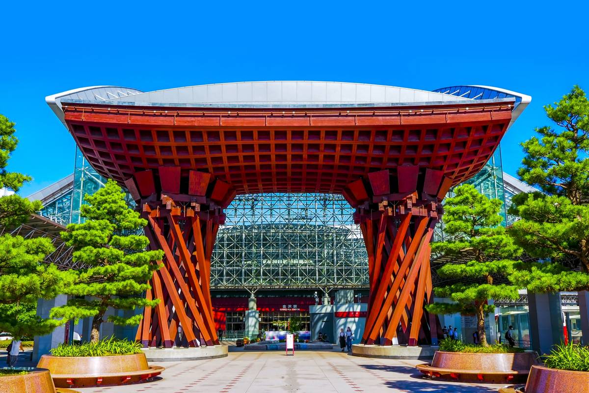 The red wooden Tsuzumi-mon Gate of Kanazawa Station, a large archway with a latticework roof, stands in front of a modern glass building.