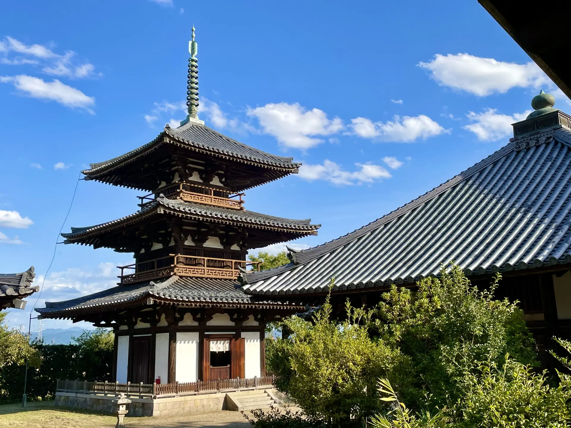 A three-tiered Japanese pagoda next to a traditional tiled-roof building under a blue sky.