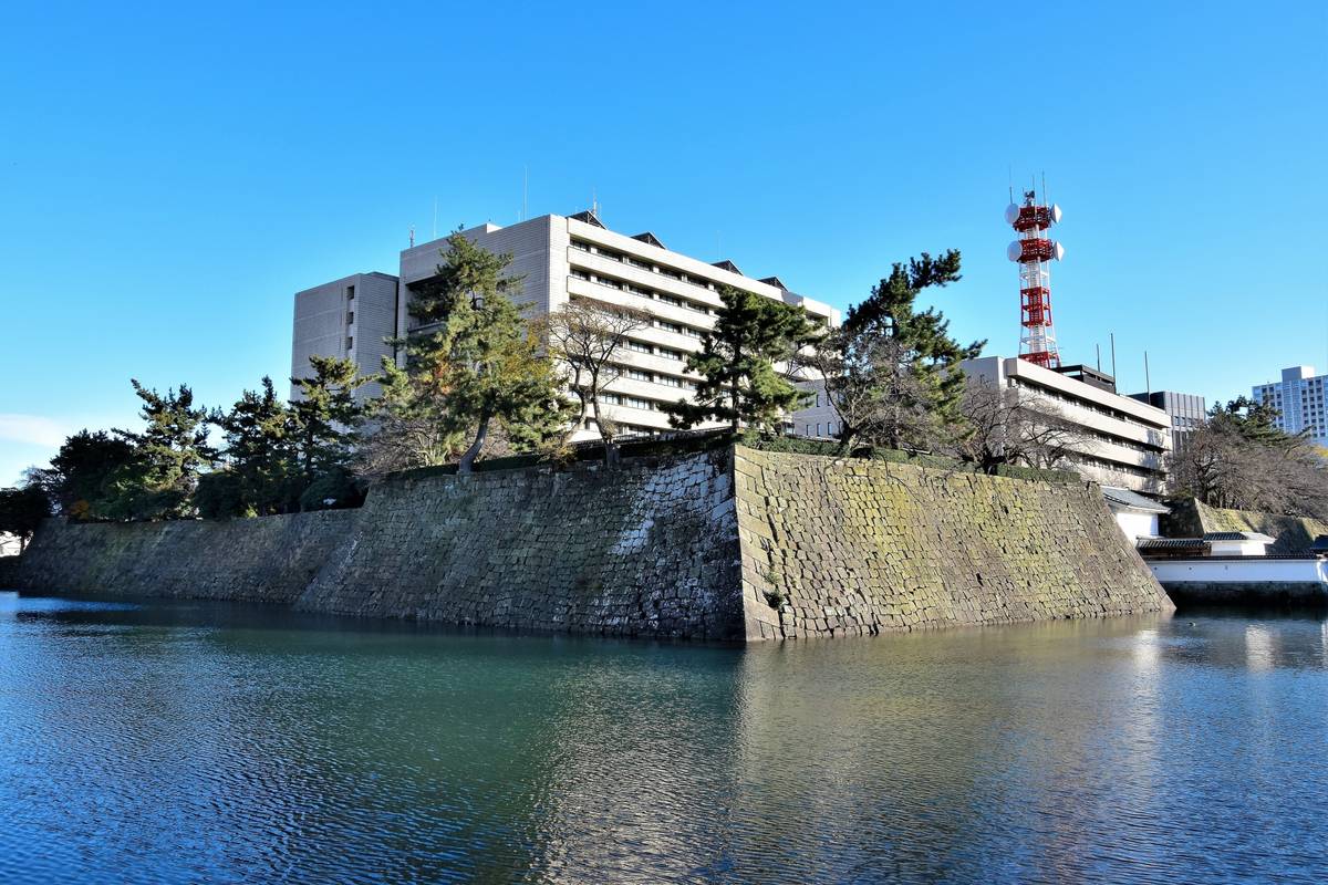 A historic stone wall and moat with modern buildings and a red and white tower in the background.