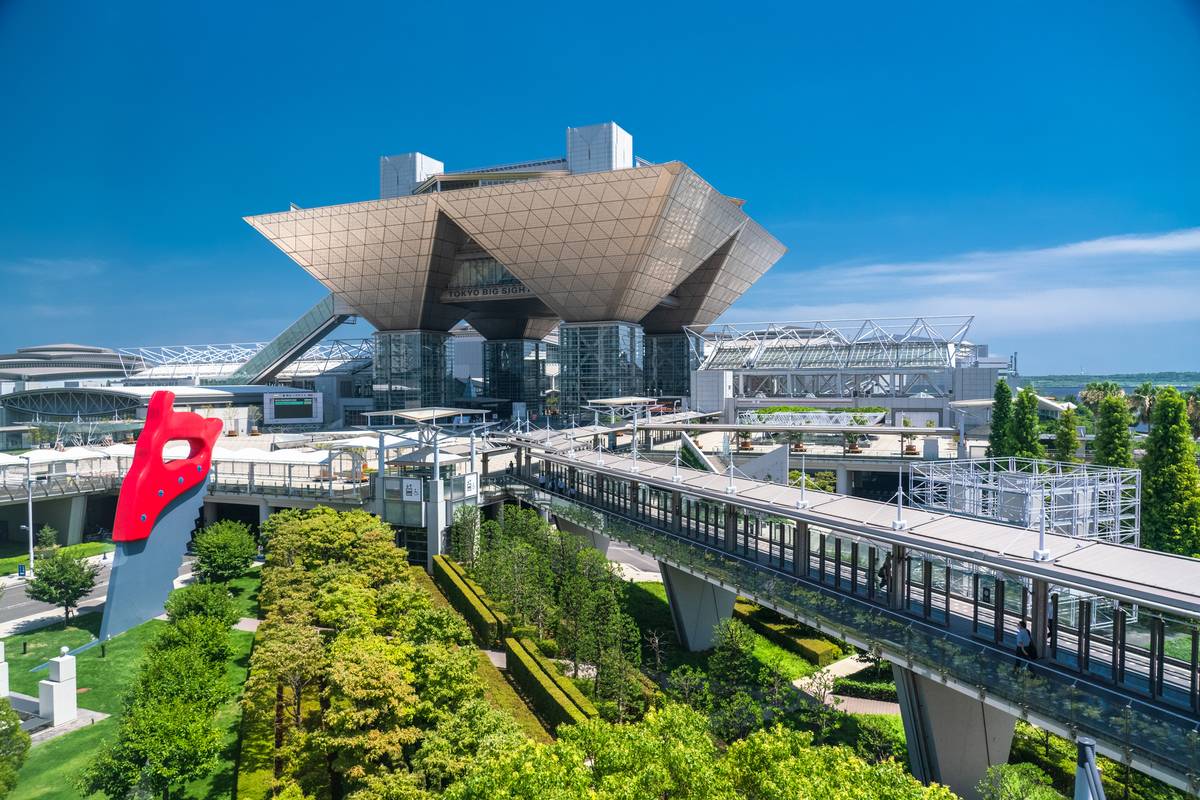Tokyo Big Sight convention center with its geometric roof, a giant red saw sculpture, and elevated walkways amidst green landscaping.