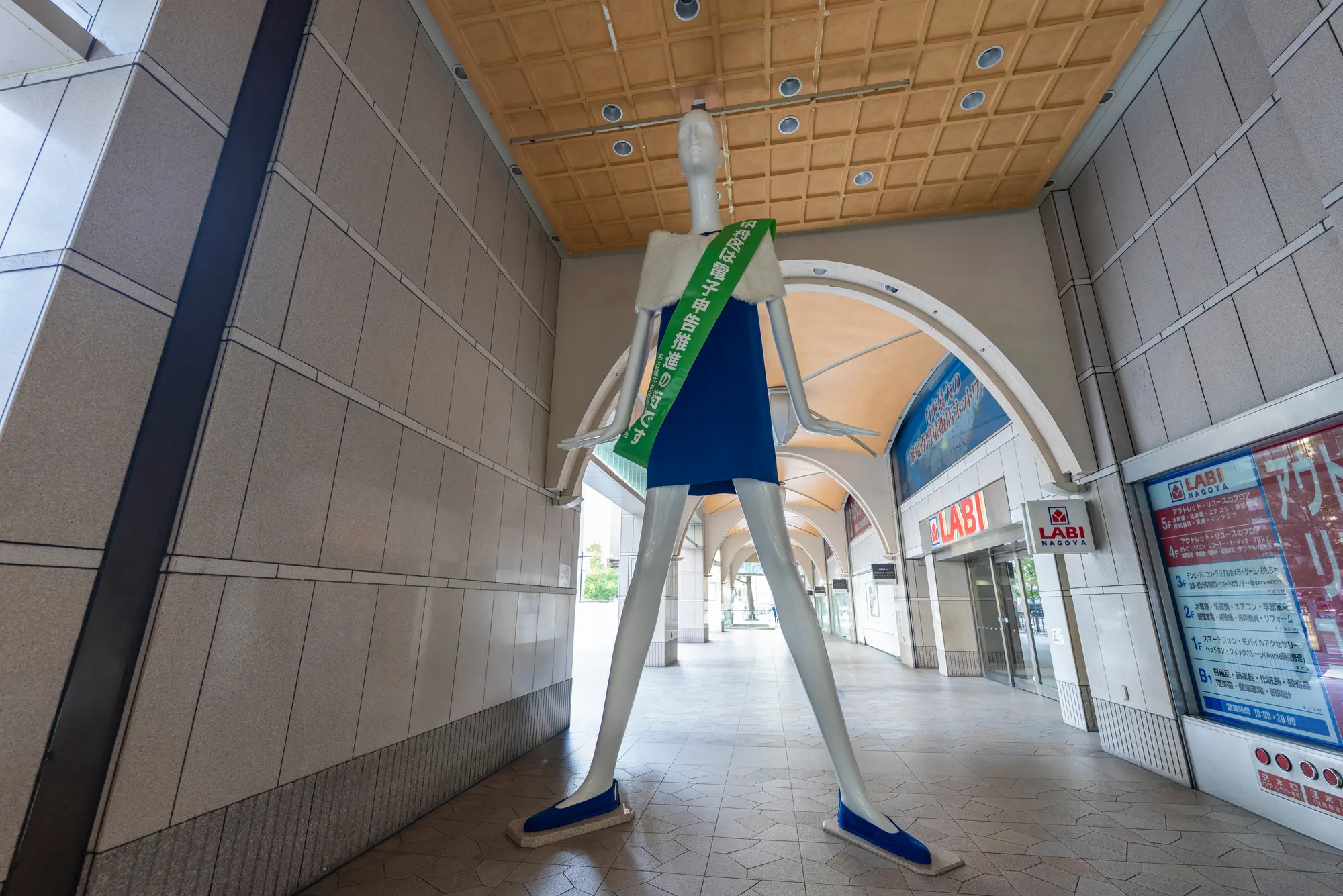 A tall, stylized mannequin wearing a blue dress and a green sash with Japanese text stands in a covered shopping arcade.