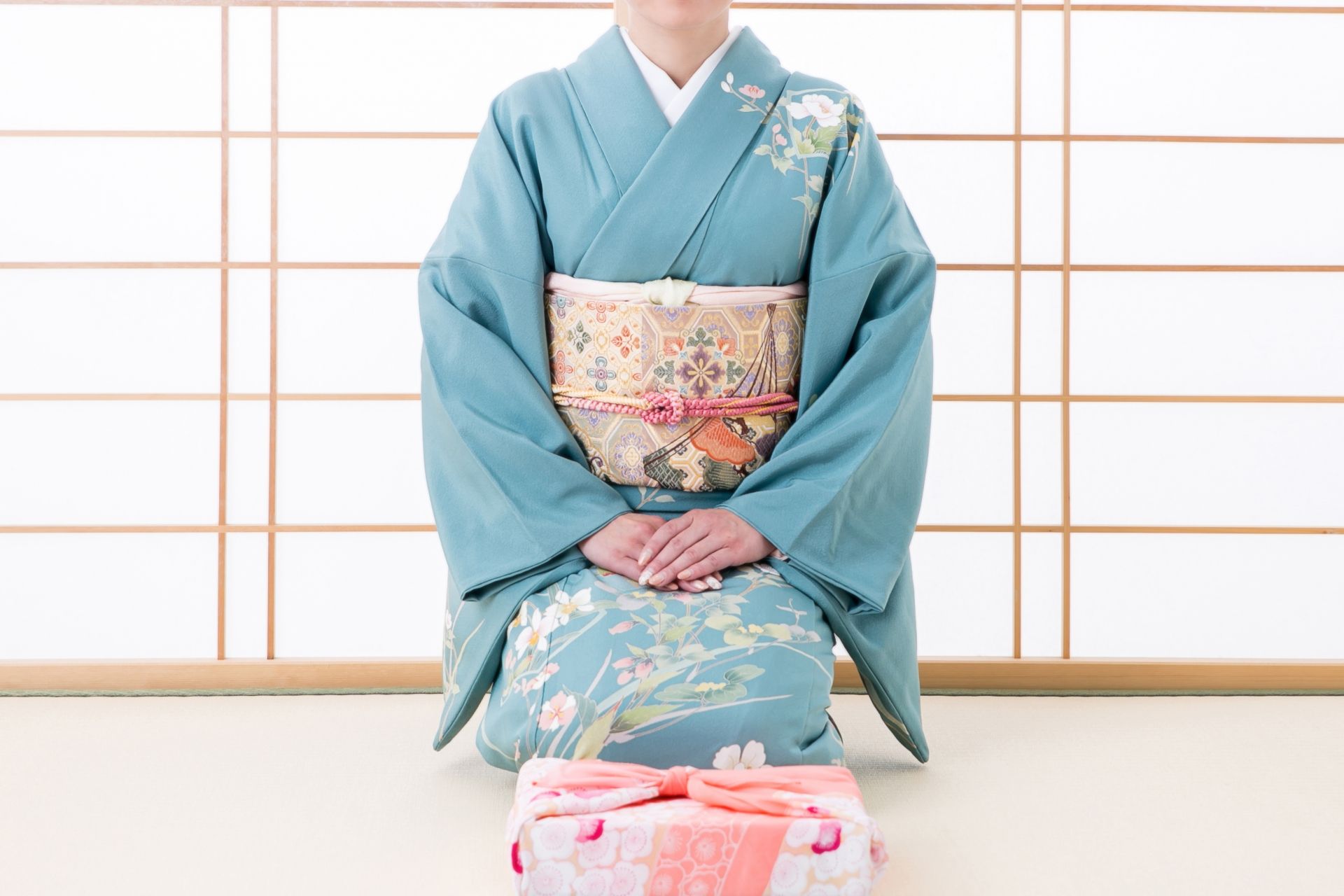 A person in a blue floral kimono kneels with a wrapped gift in front of a shoji screen.