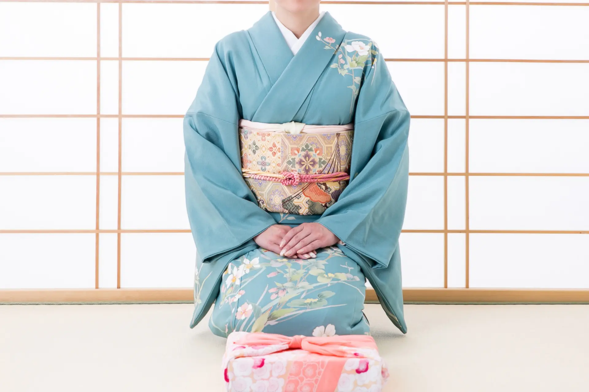 A person in a blue floral kimono kneels with a wrapped gift in front of a shoji screen.