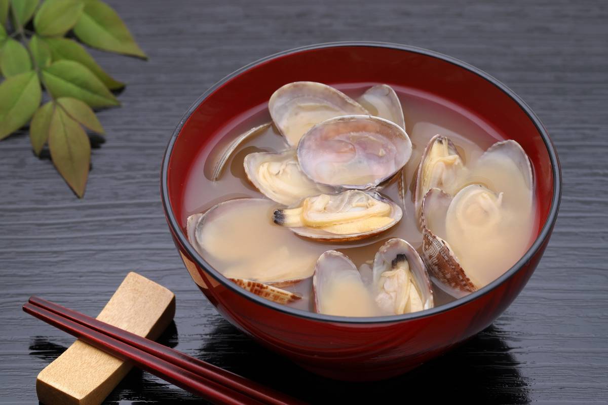 Red bowl of clam miso soup with chopsticks and green leaves on a dark table.