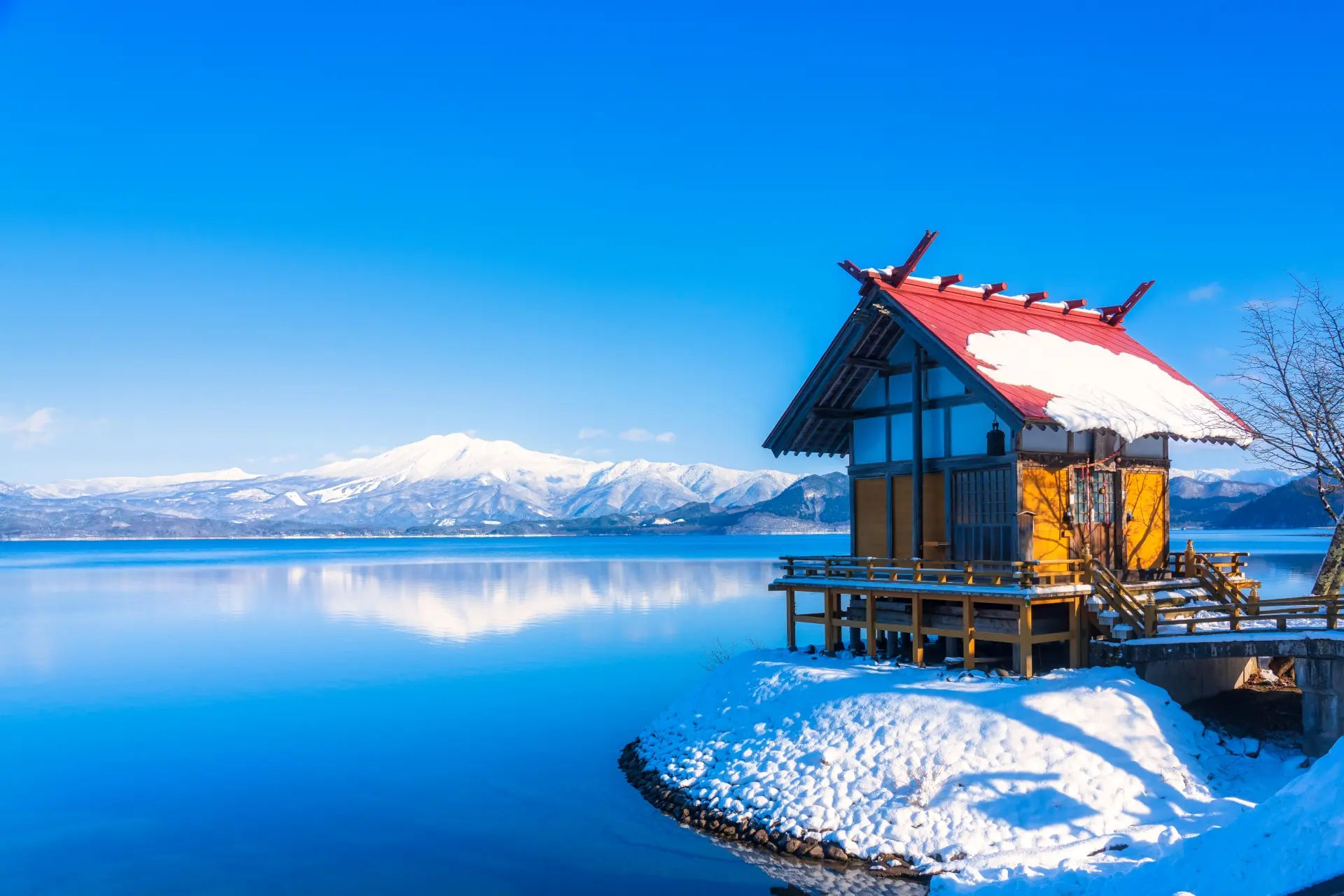 A traditional Japanese building with a red, snowy roof beside a blue lake and snow-capped mountains.