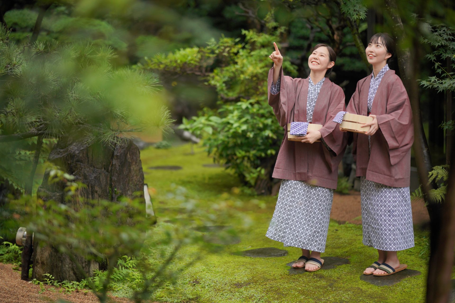 Two smiling women in yukata hold towels and wooden boxes in a lush moss garden, one pointing up.