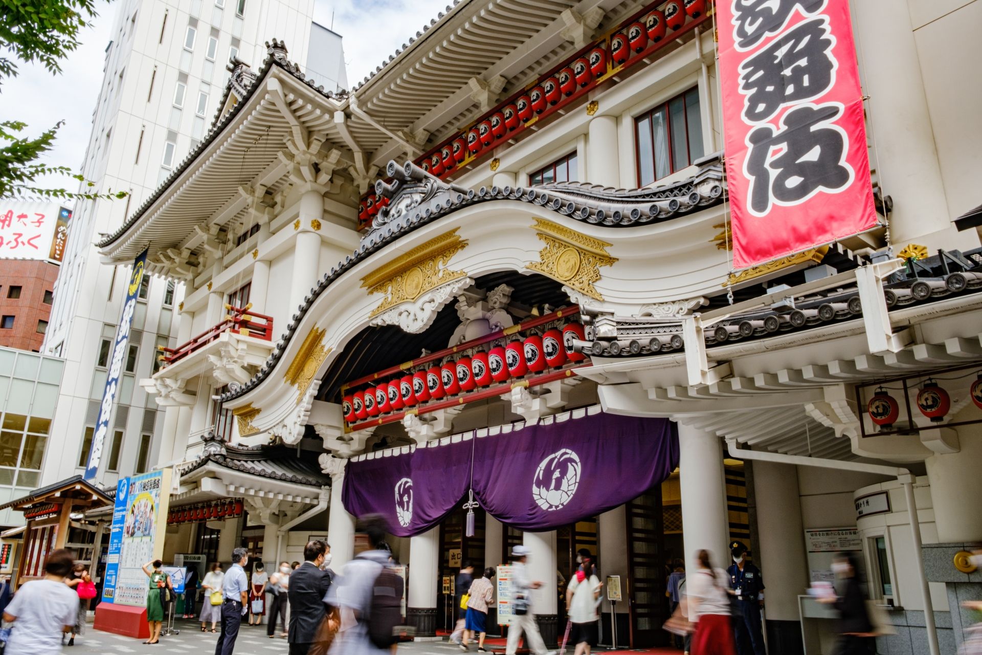 Grand exterior of a traditional Japanese Kabuki theater with red lanterns, a red banner, and people walking by.
