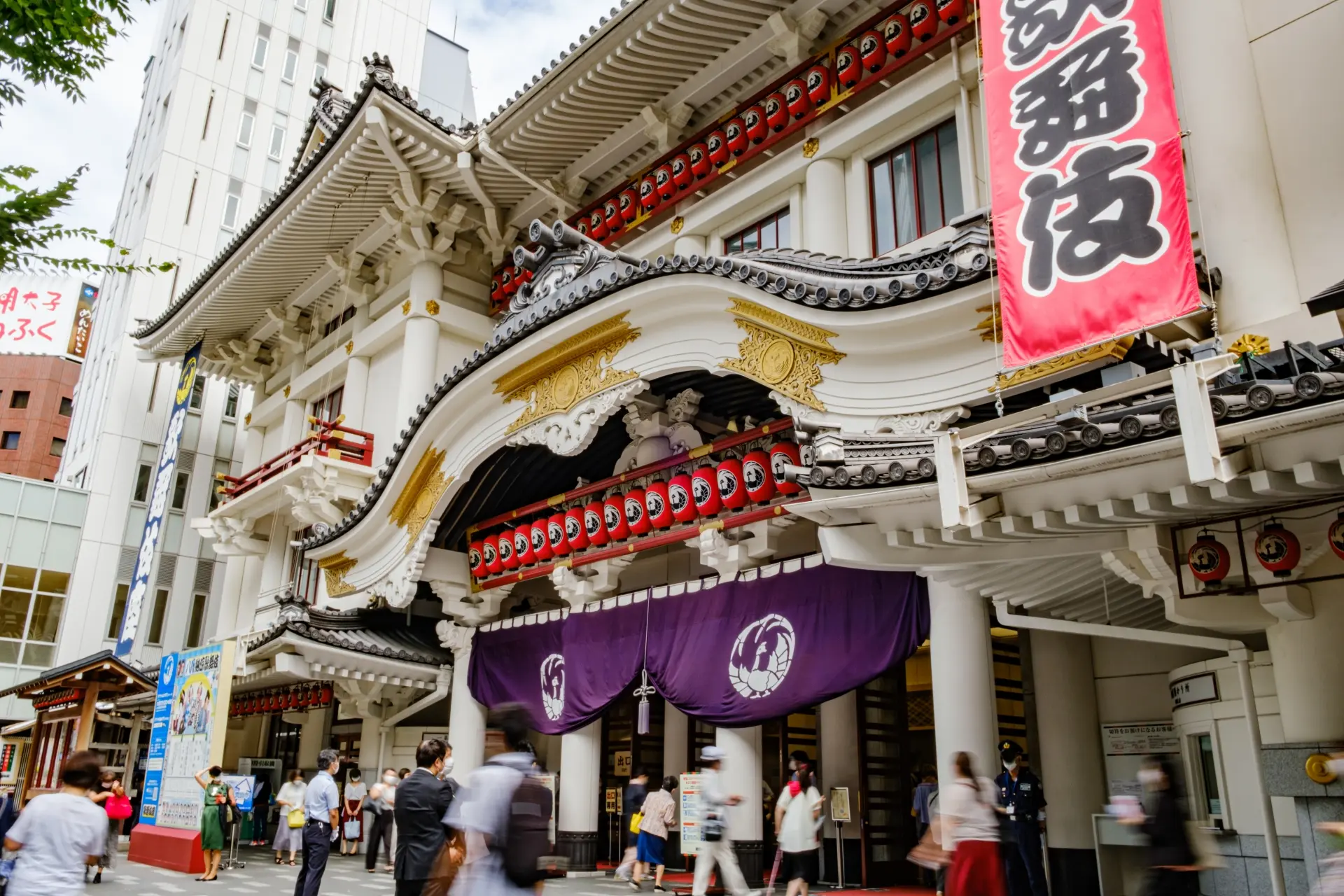 Grand exterior of a traditional Japanese Kabuki theater with red lanterns, a red banner, and people walking by.