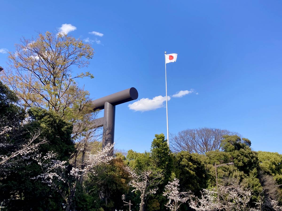 Japanese flag flying near a large torii gate amidst trees with cherry blossoms under a blue sky.