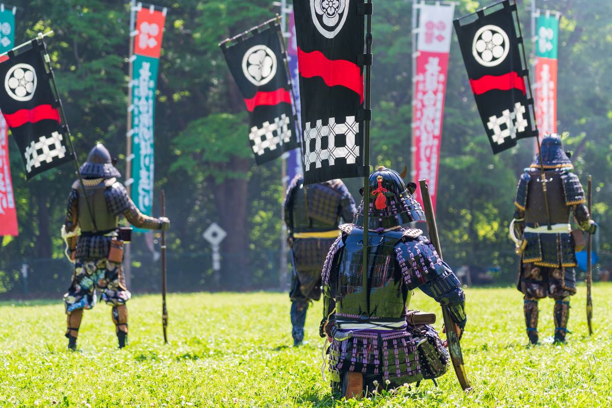 Several people in samurai armor stand in a grassy field surrounded by traditional Japanese flags.