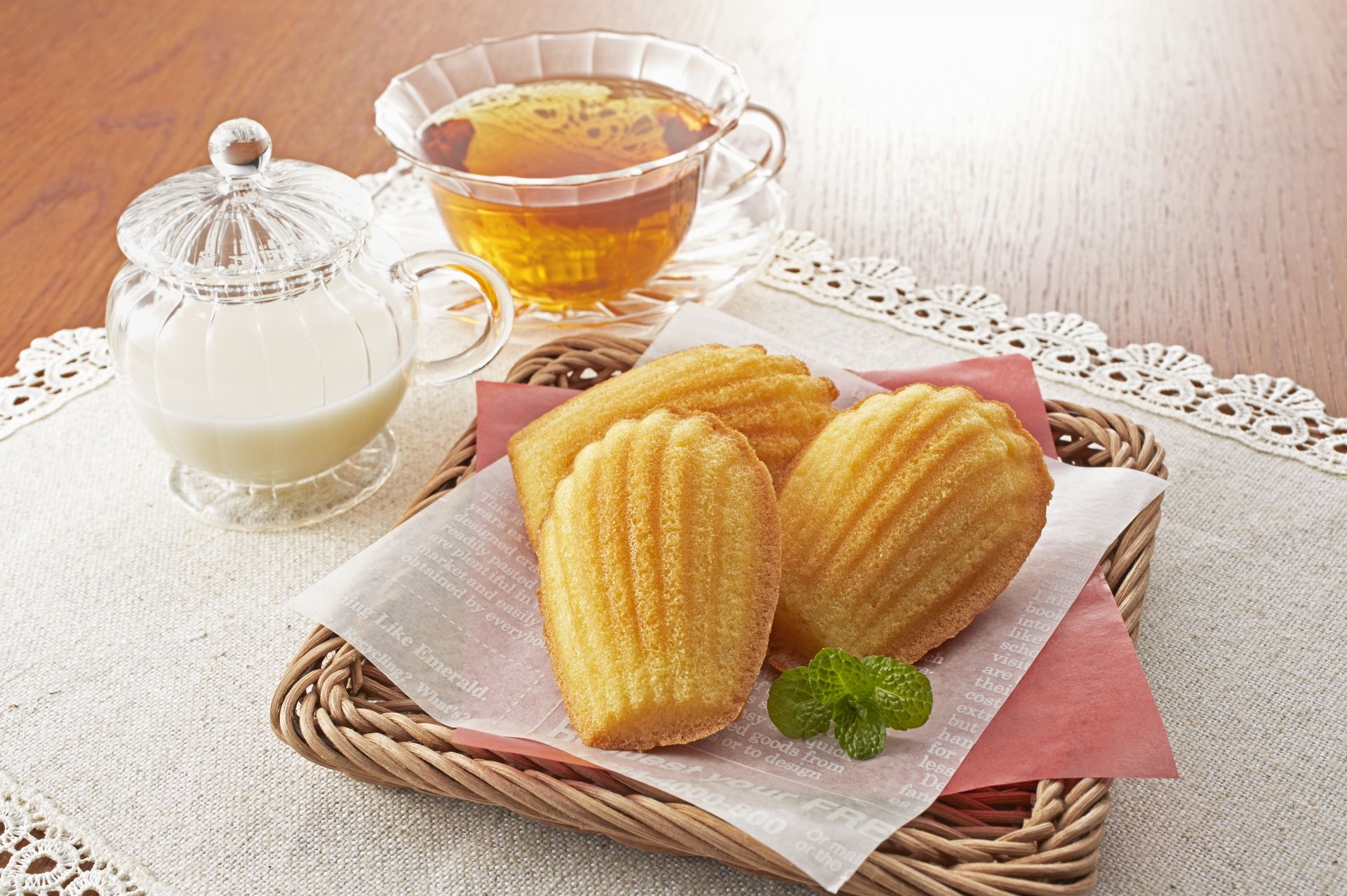 Four golden madeleines in a wicker basket with a sprig of mint, alongside a cup of tea and a glass creamer of milk