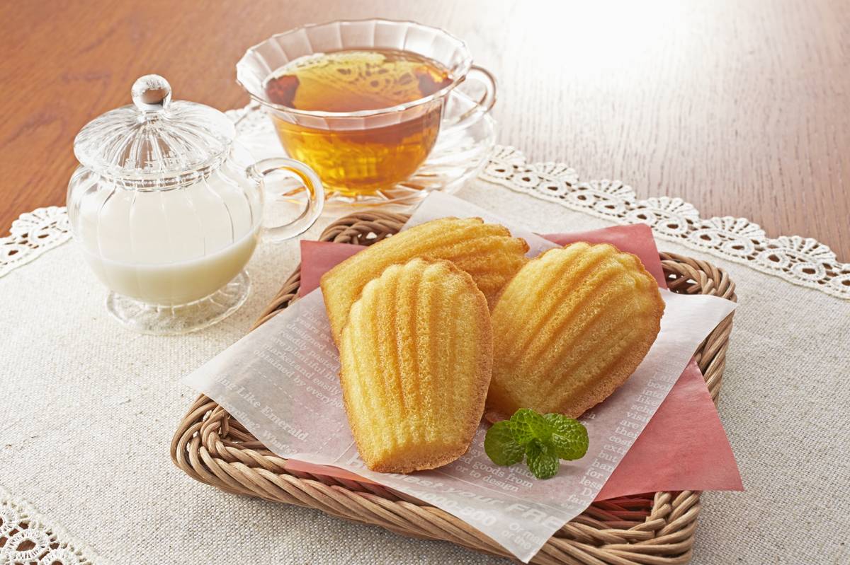 Four golden madeleines in a wicker basket with a sprig of mint, alongside a cup of tea and a glass creamer of milk
