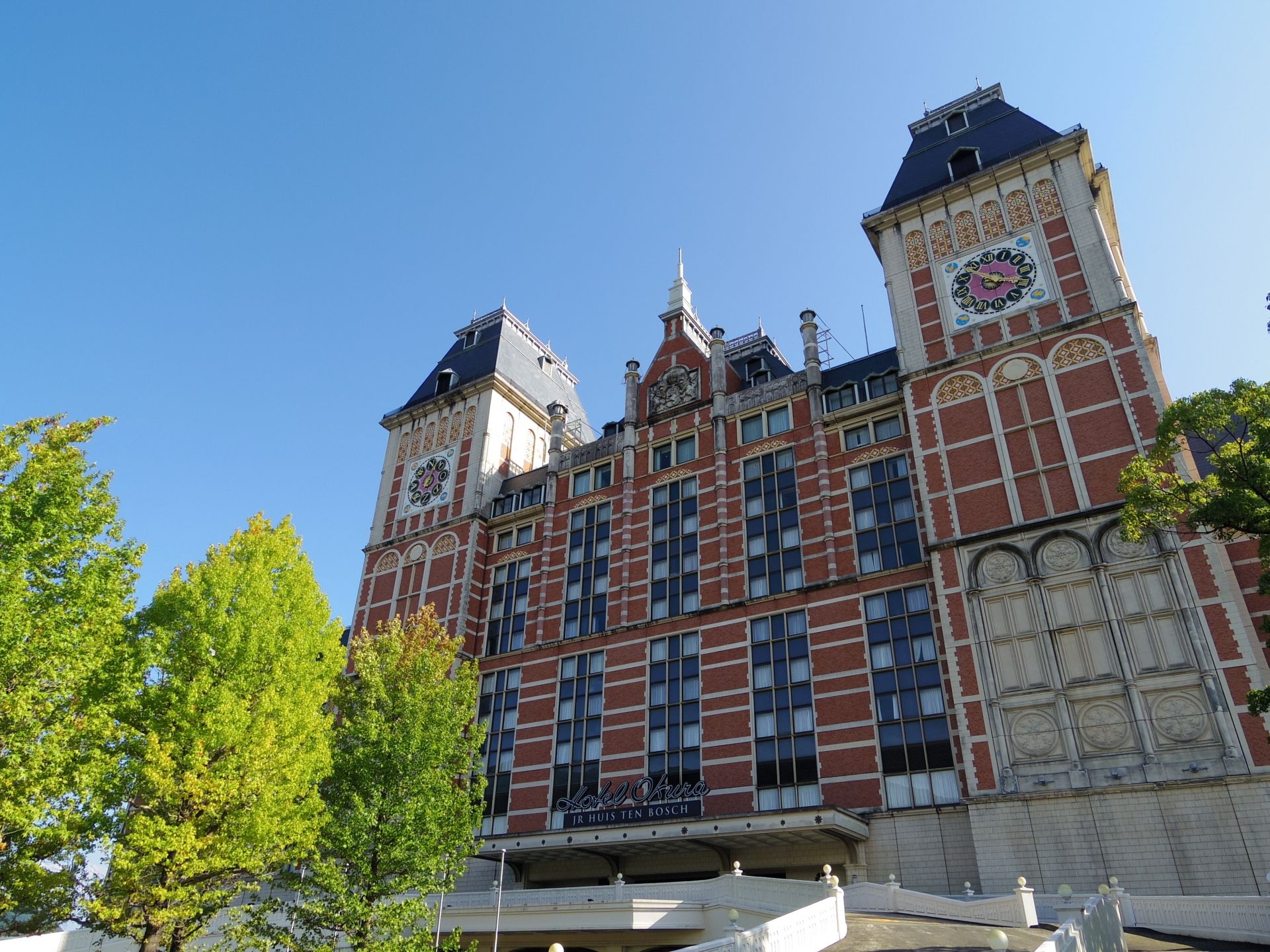 A large, ornate red brick building with two clock towers, framed by green trees against a clear blue sky.