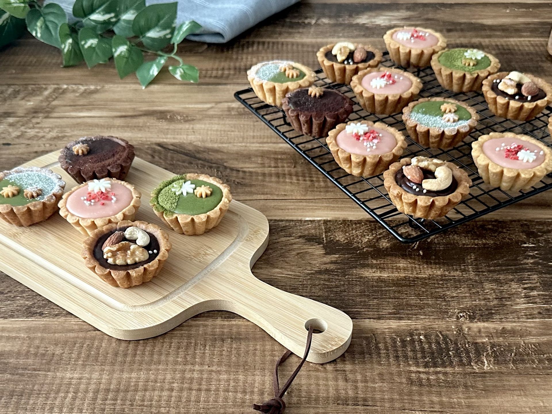 An assortment of colorful mini tarts with various toppings displayed on a wooden board and a cooling rack.