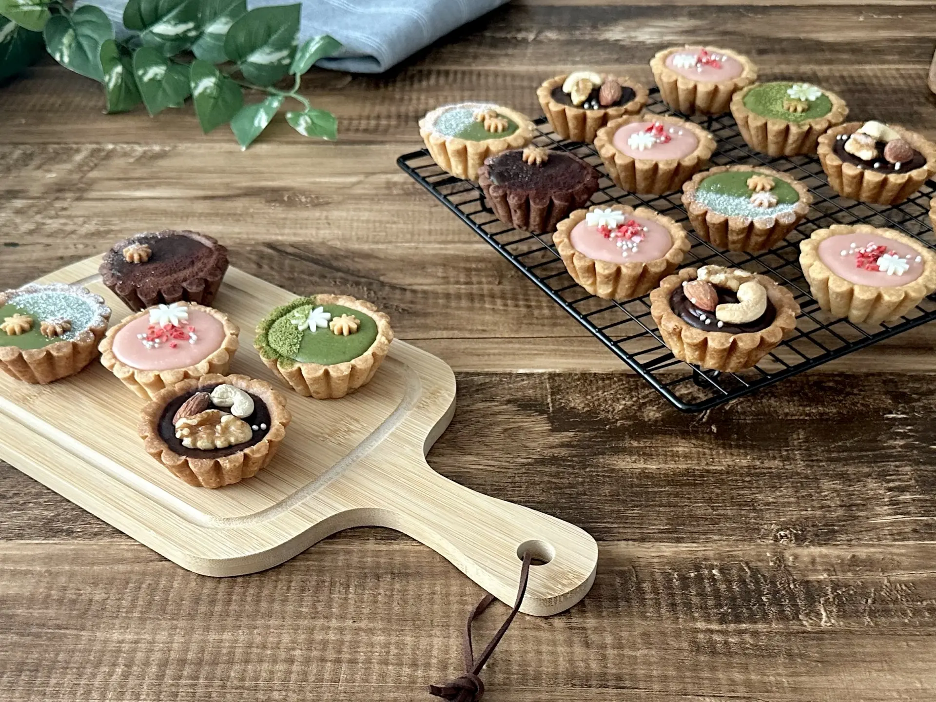 An assortment of colorful mini tarts with various toppings displayed on a wooden board and a cooling rack.
