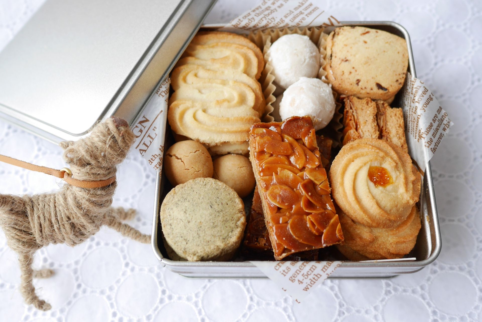 An open tin filled with assorted cookies and baked goods, next to a small twine dog on a white lace tablecloth.