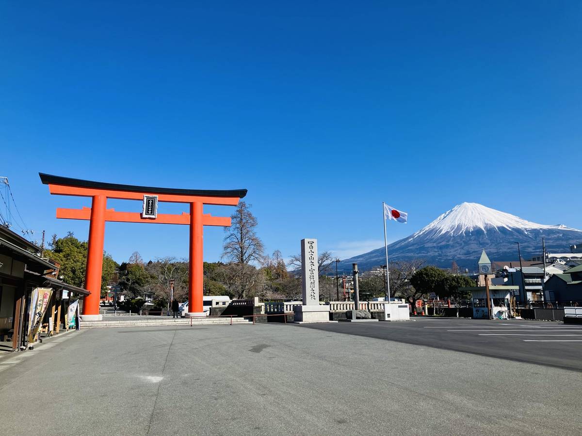 A large orange torii gate stands before snow-capped Mount Fuji and a Japanese flag under a clear blue sky.