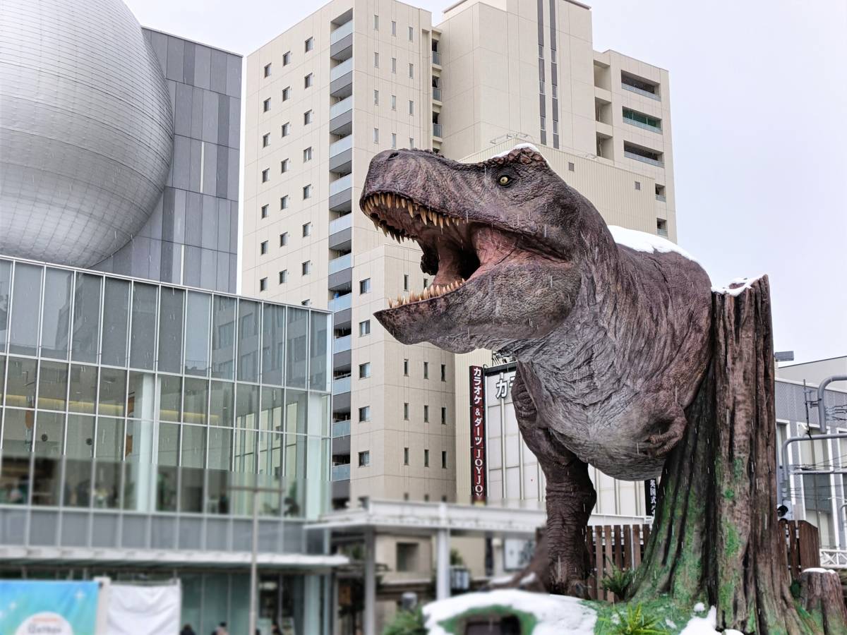 A T-Rex statue emerging from a tree stump, lightly covered in snow, against a backdrop of modern city buildings.