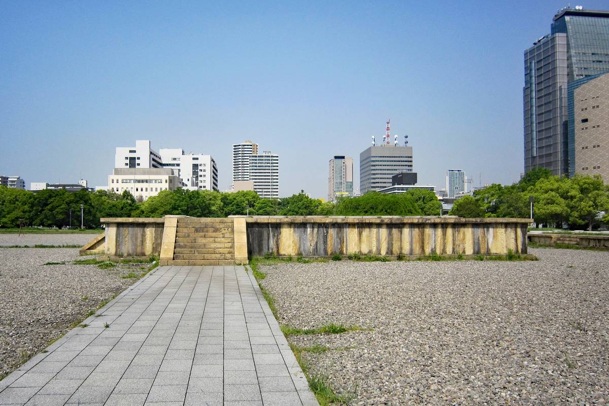 A paved path through gravel leads to a low wall with steps, backed by a city skyline and trees.