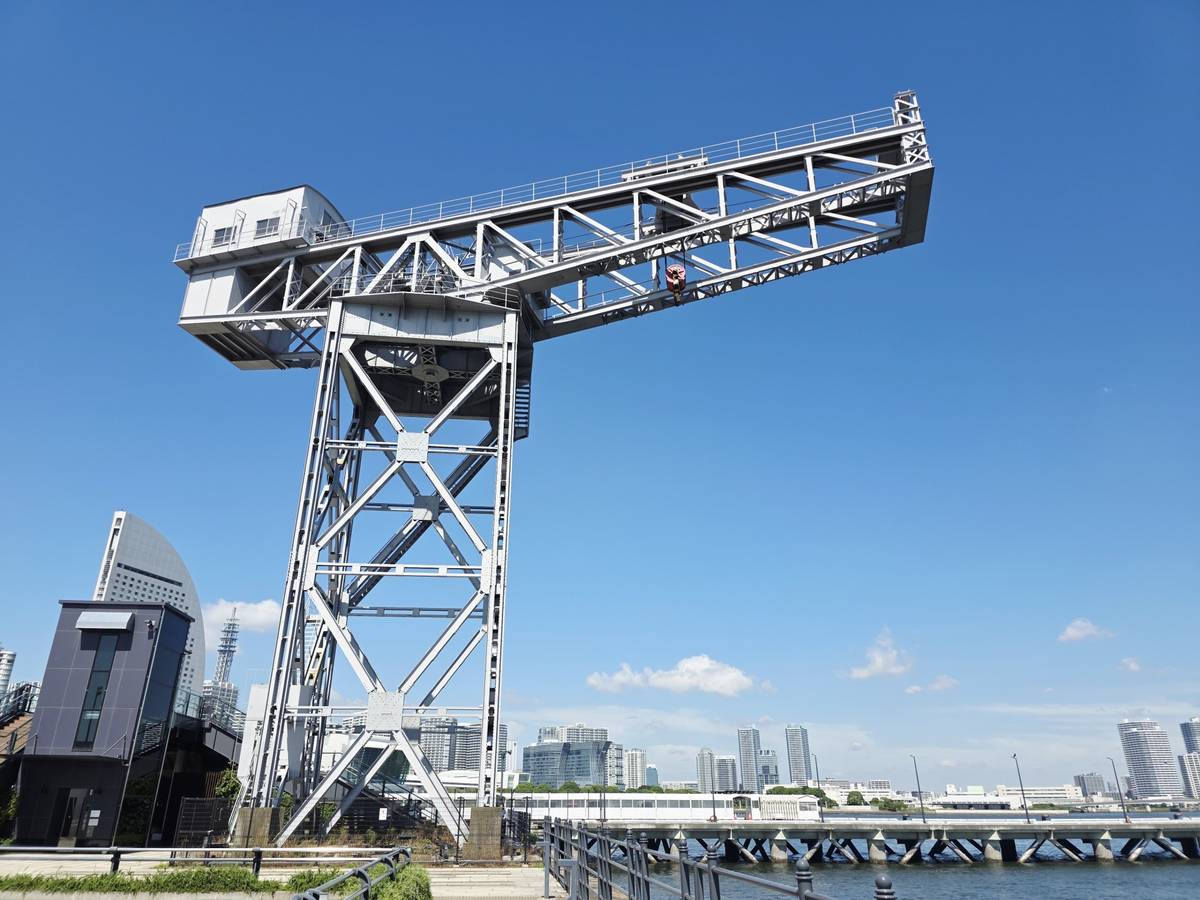 A large, grey industrial crane stands tall against a clear blue sky, overlooking a city waterfront with buildings in the distance.