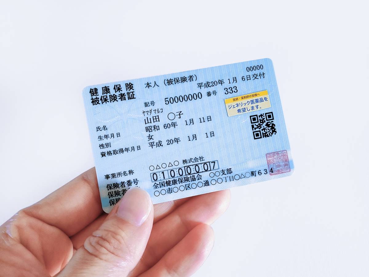 A hand holds a light blue Japanese health insurance card with text, numbers, and a QR code.