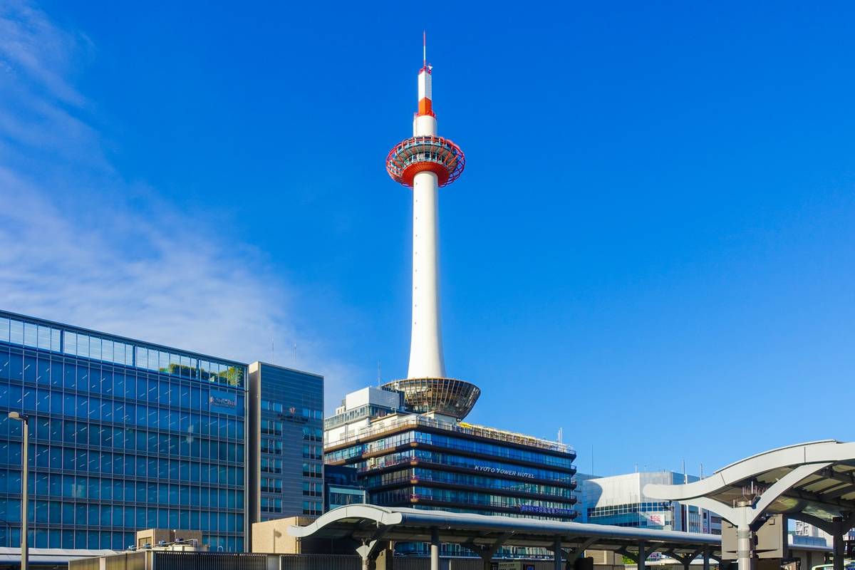 Kyoto Tower, a white and red observation tower, stands tall against a clear blue sky, flanked by modern glass buildings.