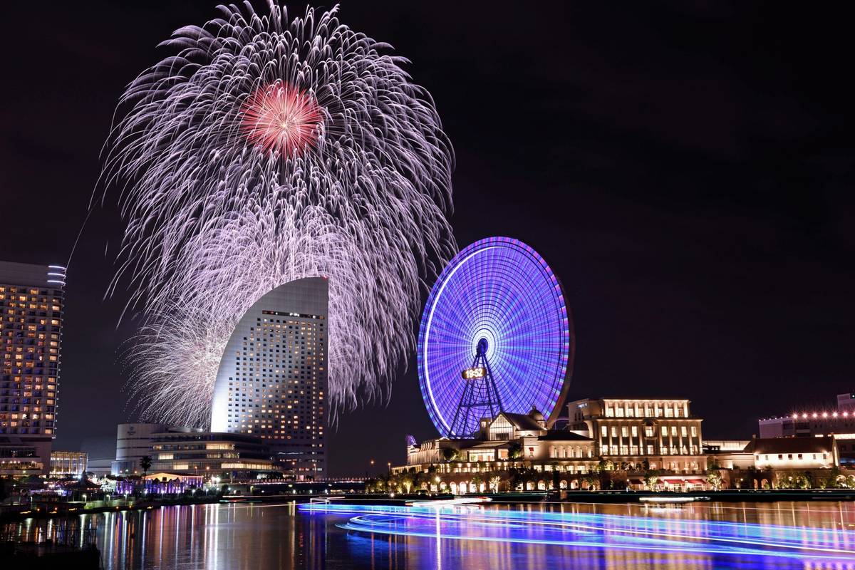Fireworks explode over a city skyline at night, with a purple-lit Ferris wheel and light trails on the water.