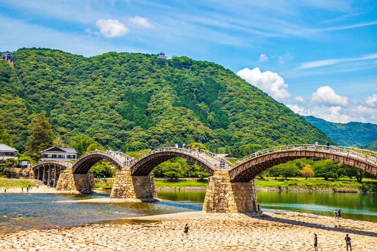 Kintaikyo Bridge, a multi-arched wooden bridge, spans a river with lush green mountains and Iwakuni Castle in the background.