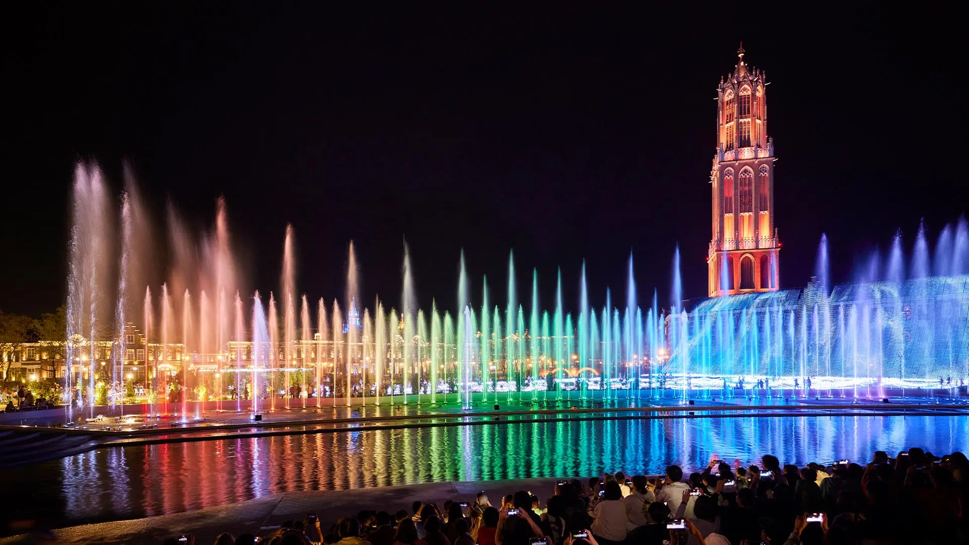 Colorful night fountain show with a tall illuminated tower and its reflection, viewed by a crowd.
