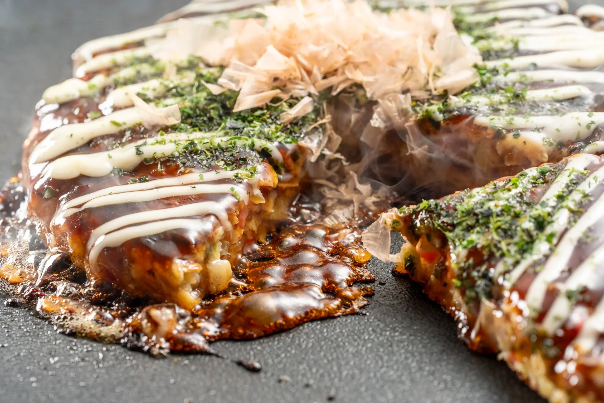 A steaming okonomiyaki, sliced and topped with dark sauce, mayonnaise, green flakes, and bonito flakes.