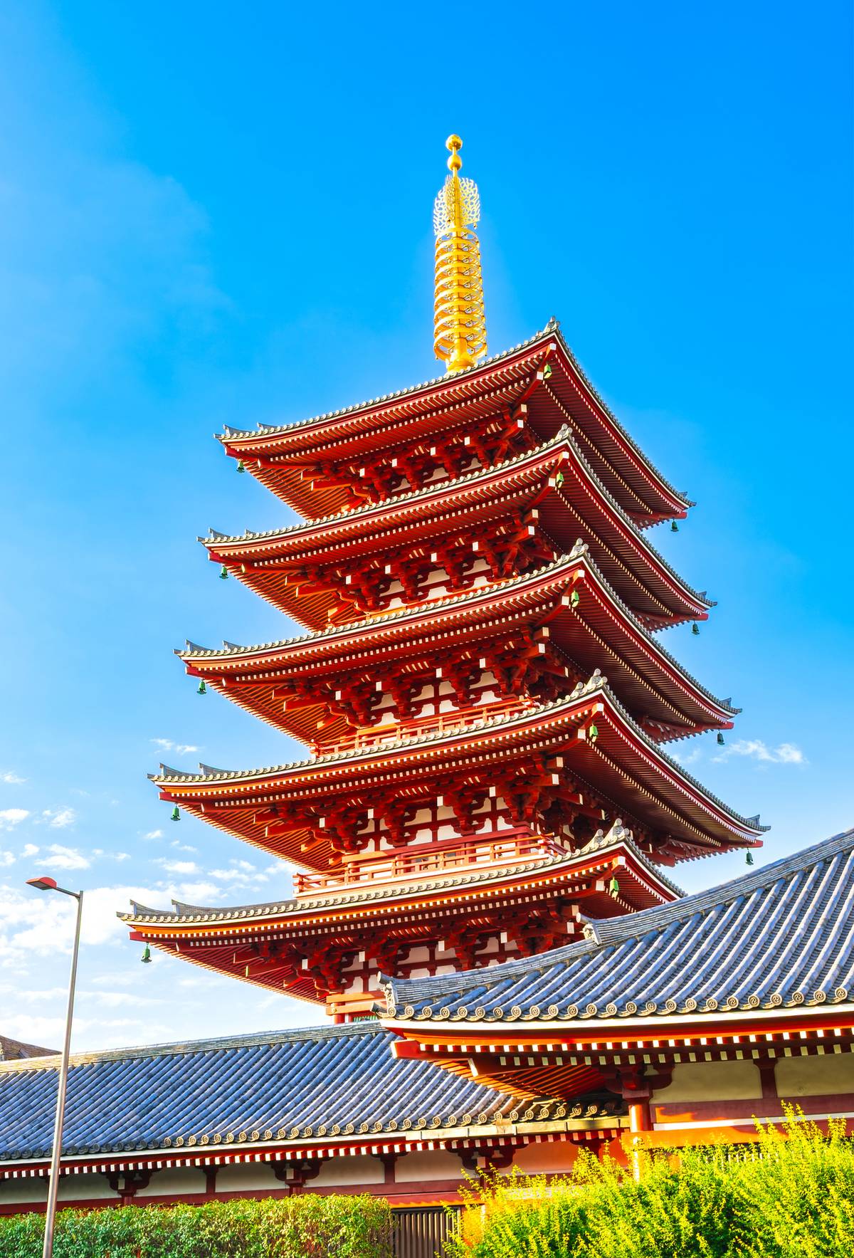 A tall, red five-story pagoda with a golden spire stands against a clear blue sky, with parts of dark-tiled temple roofs and green foliage visible in the foreground.