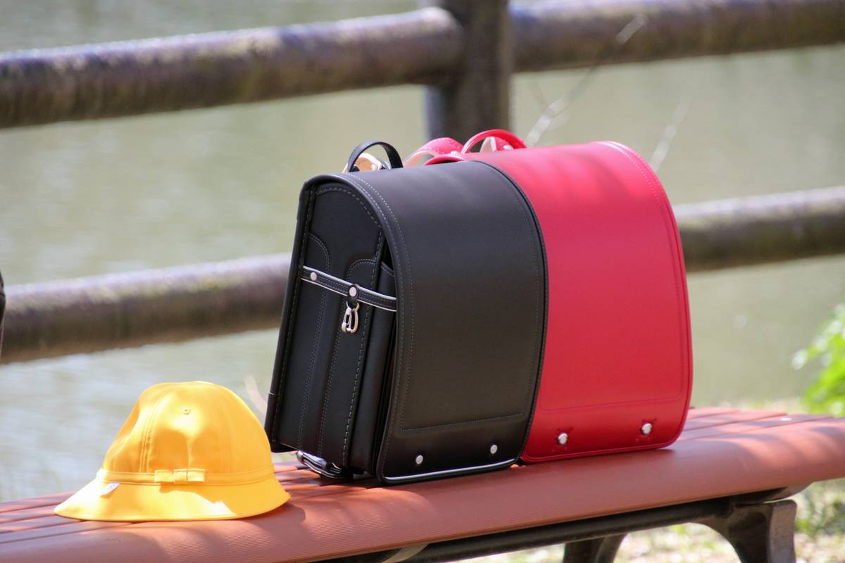 A black and red randoseru and a yellow hat rest on a brown bench.