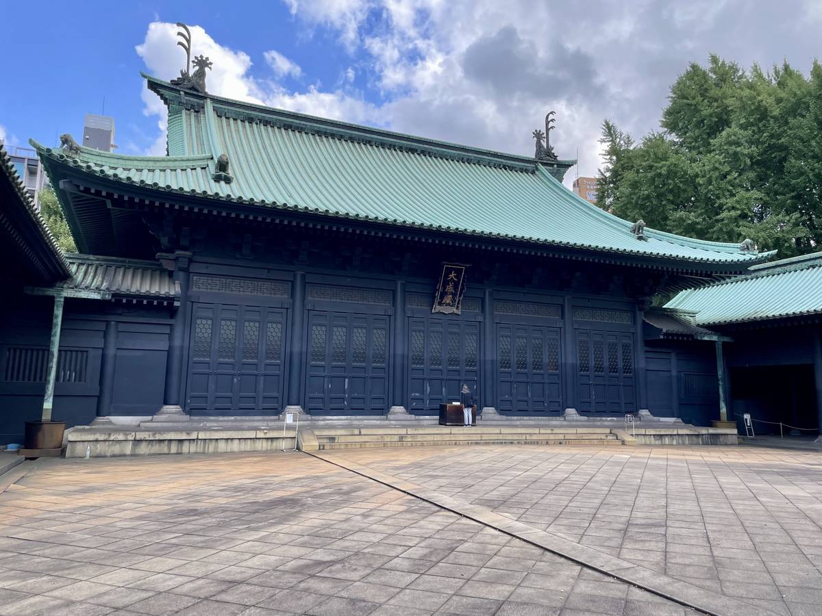 Dark traditional Japanese temple with a curved green roof, a sign over its main doors, and a paved courtyard.