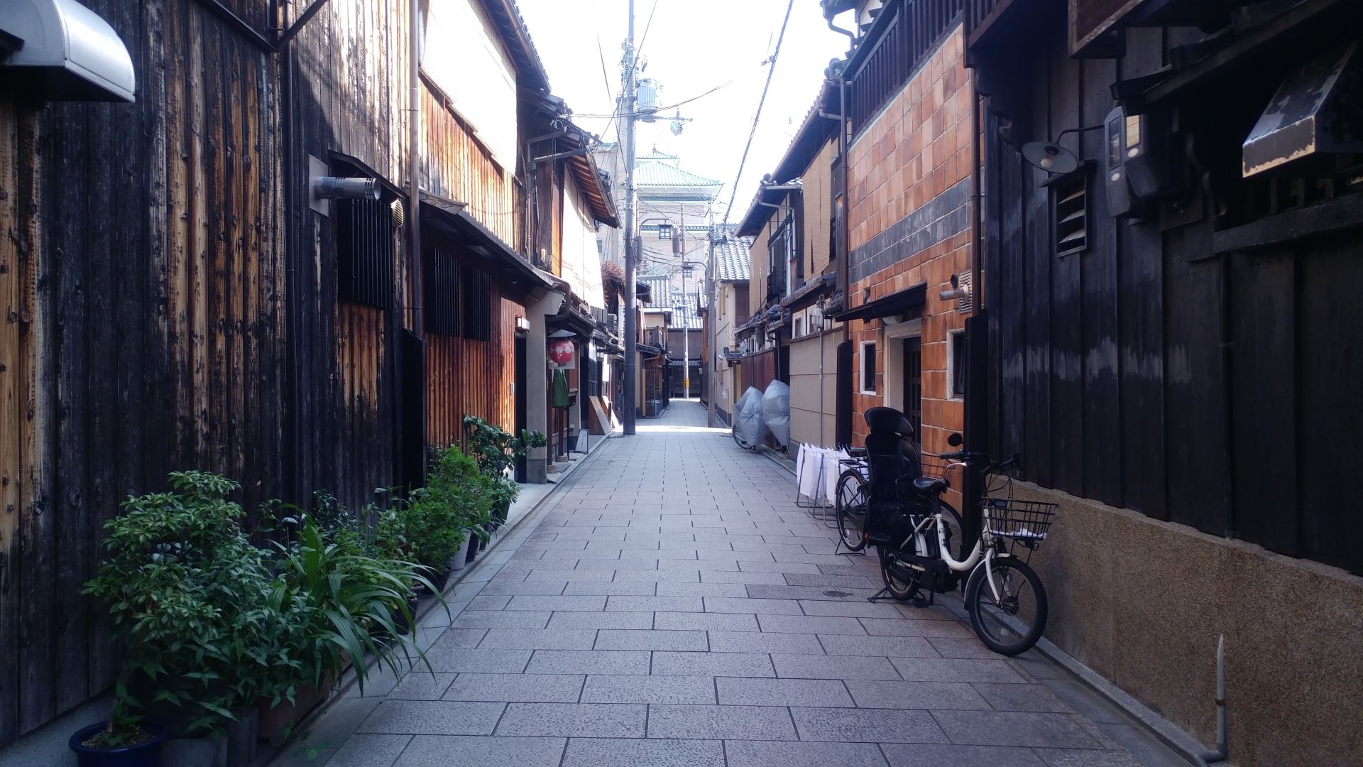 A narrow, paved alley lined with traditional wooden buildings, potted plants, and a bicycle.
