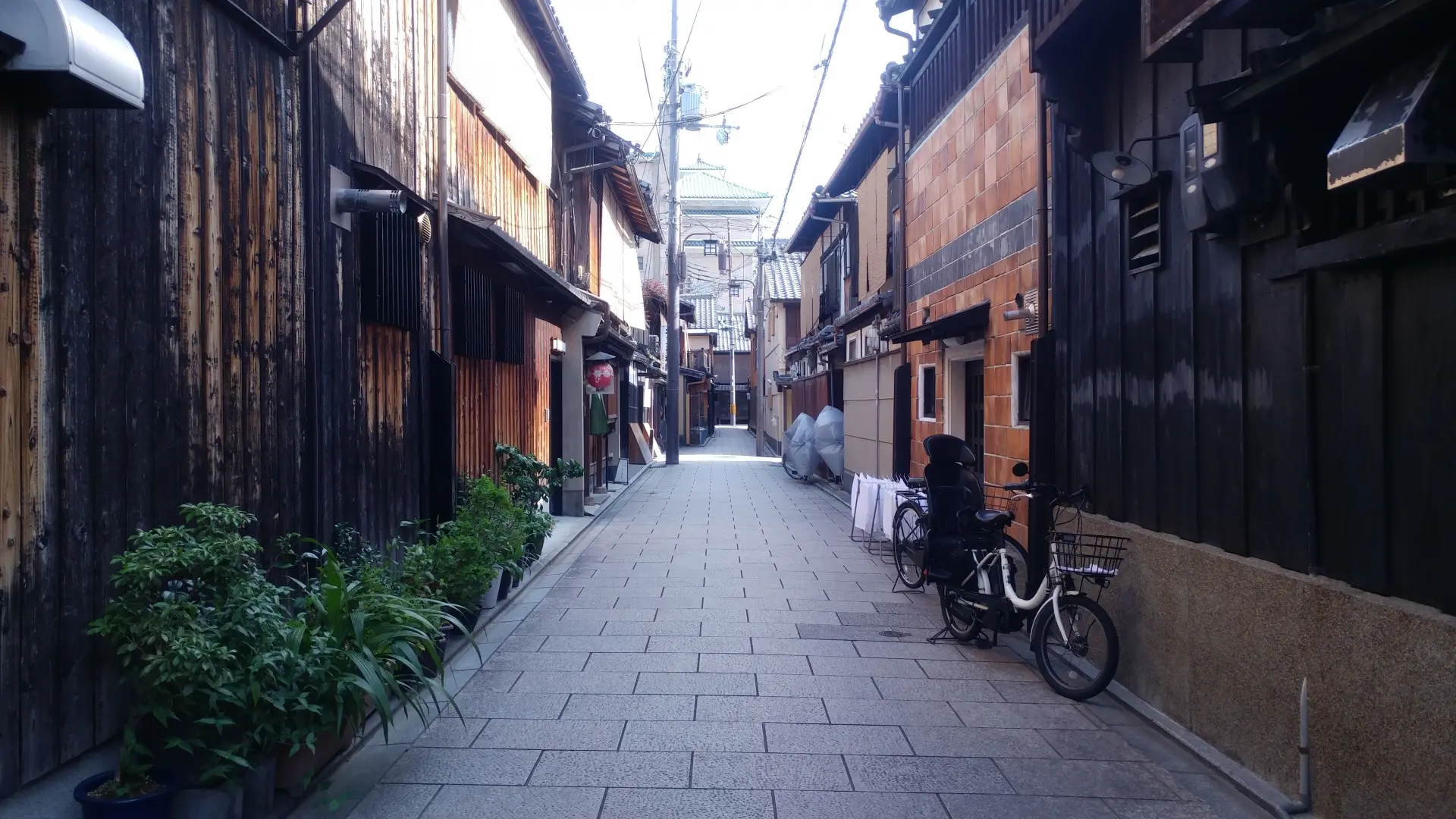 A narrow, paved alley lined with traditional wooden buildings, potted plants, and a bicycle.