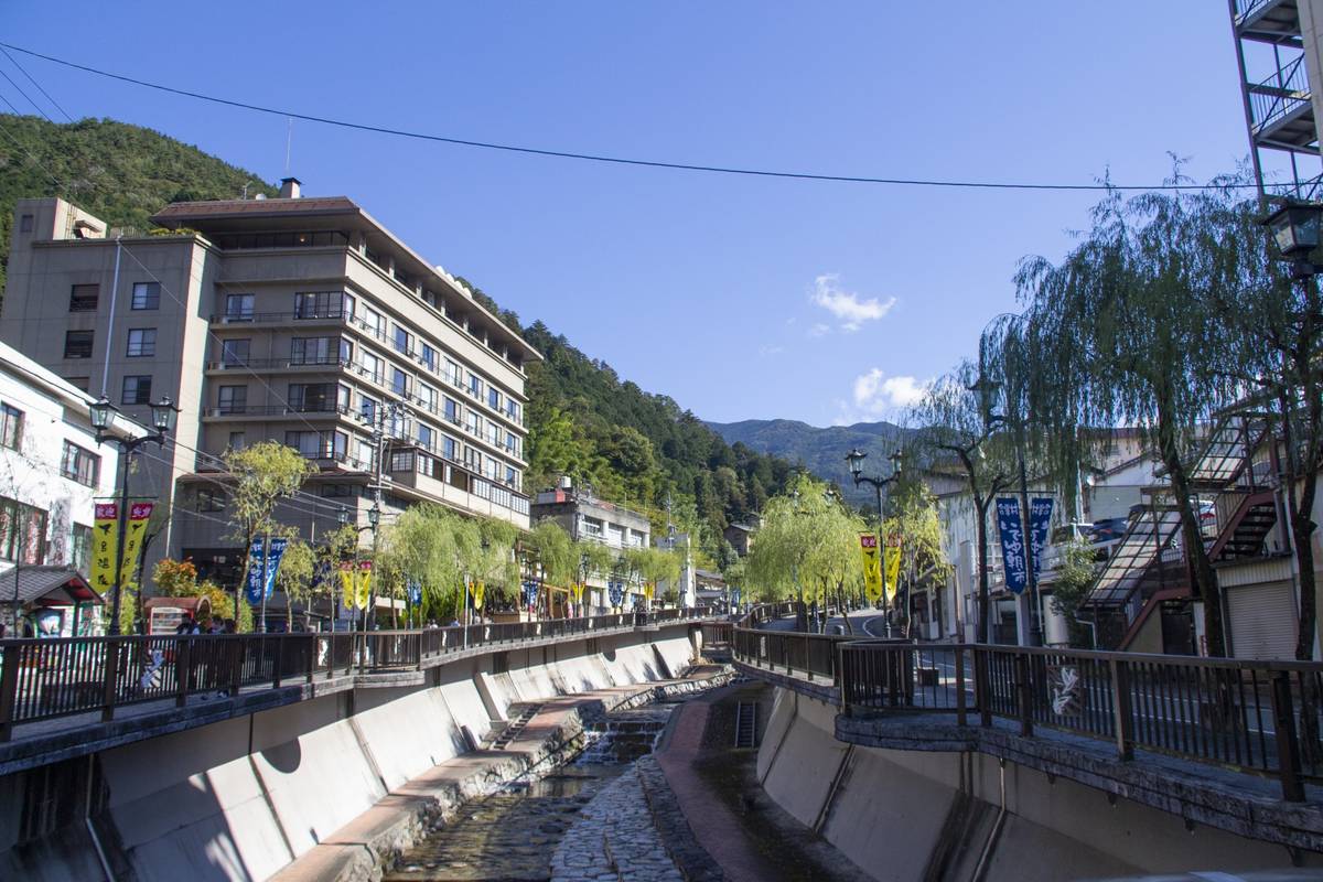 A river flows through a town lined with buildings and walkways, with green mountains in the background under a blue sky.