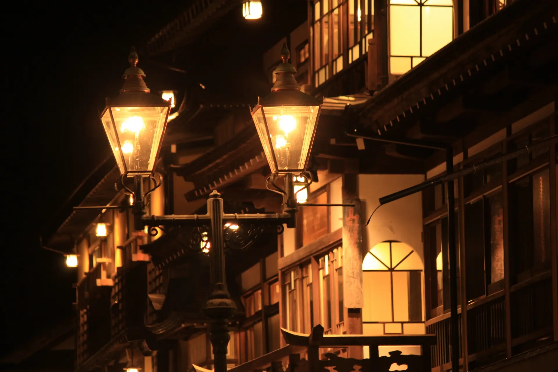 Two vintage streetlights glow in front of traditional buildings at night.