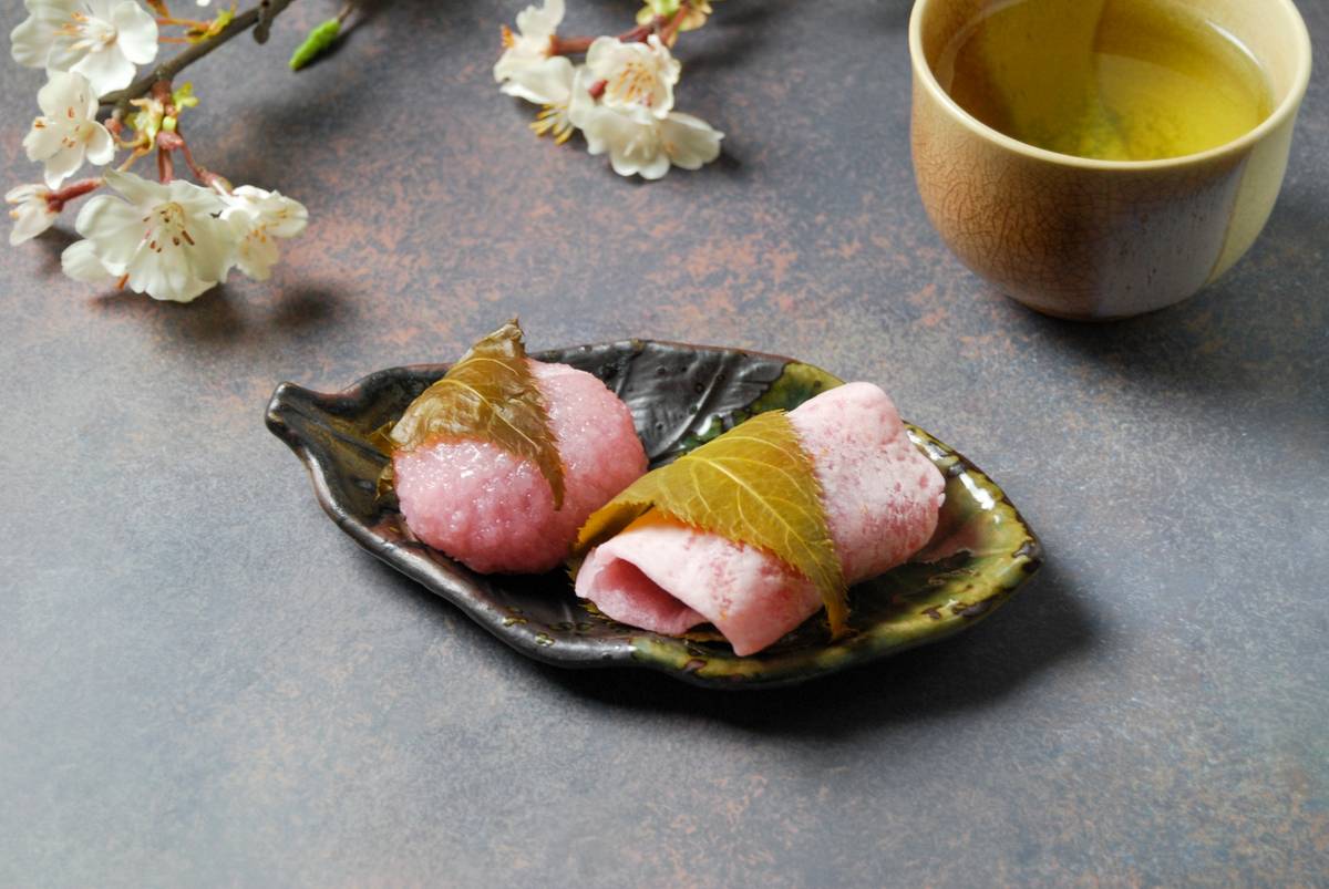 Two pink sakura mochi on a plate with green tea and cherry blossoms.