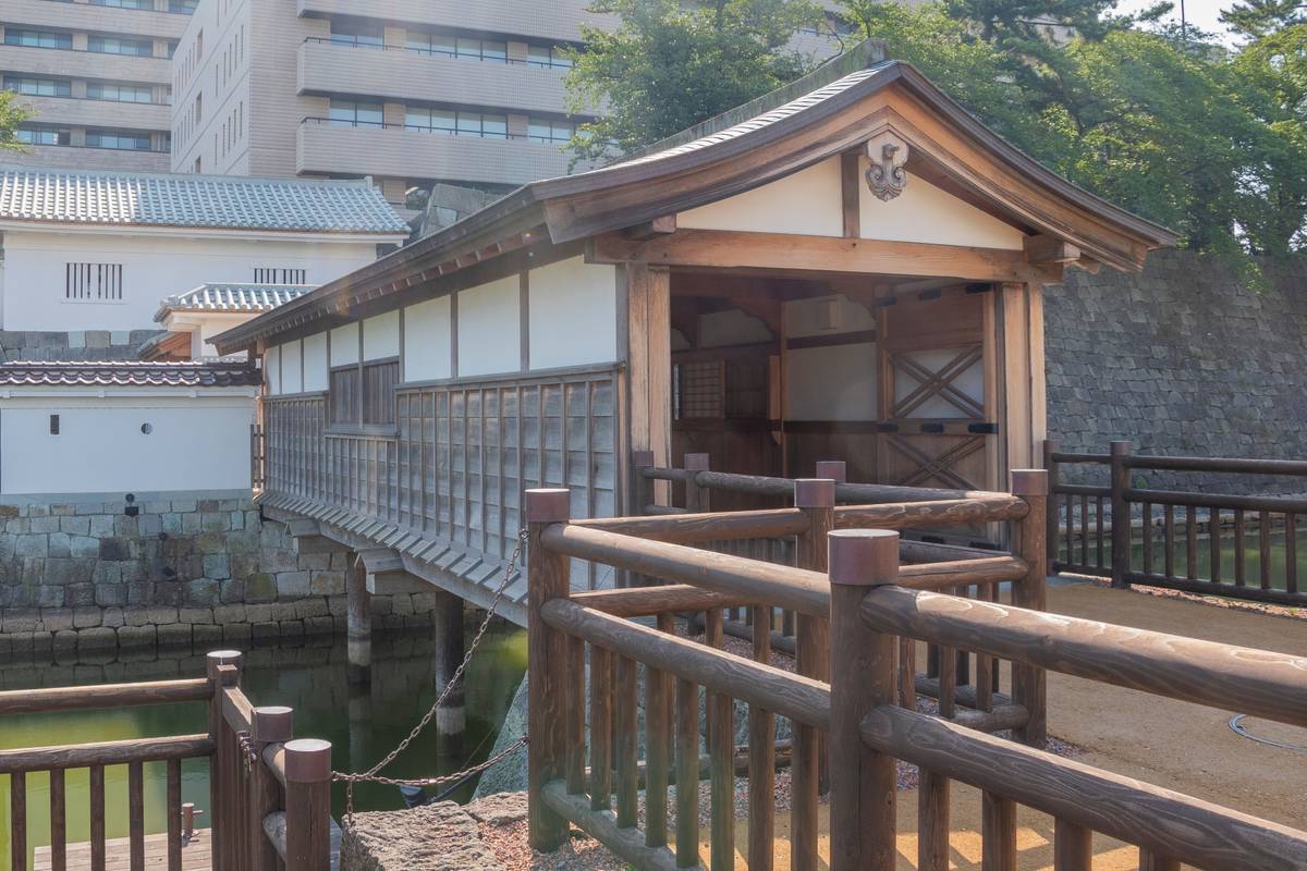 Traditional Japanese covered wooden bridge over a moat, next to castle walls.