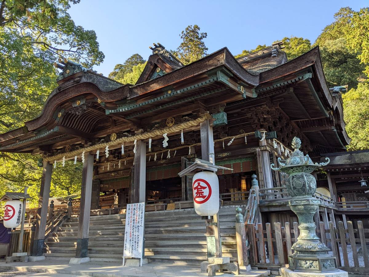 A traditional Japanese wooden shrine with a curved roof, lanterns, and green trees.