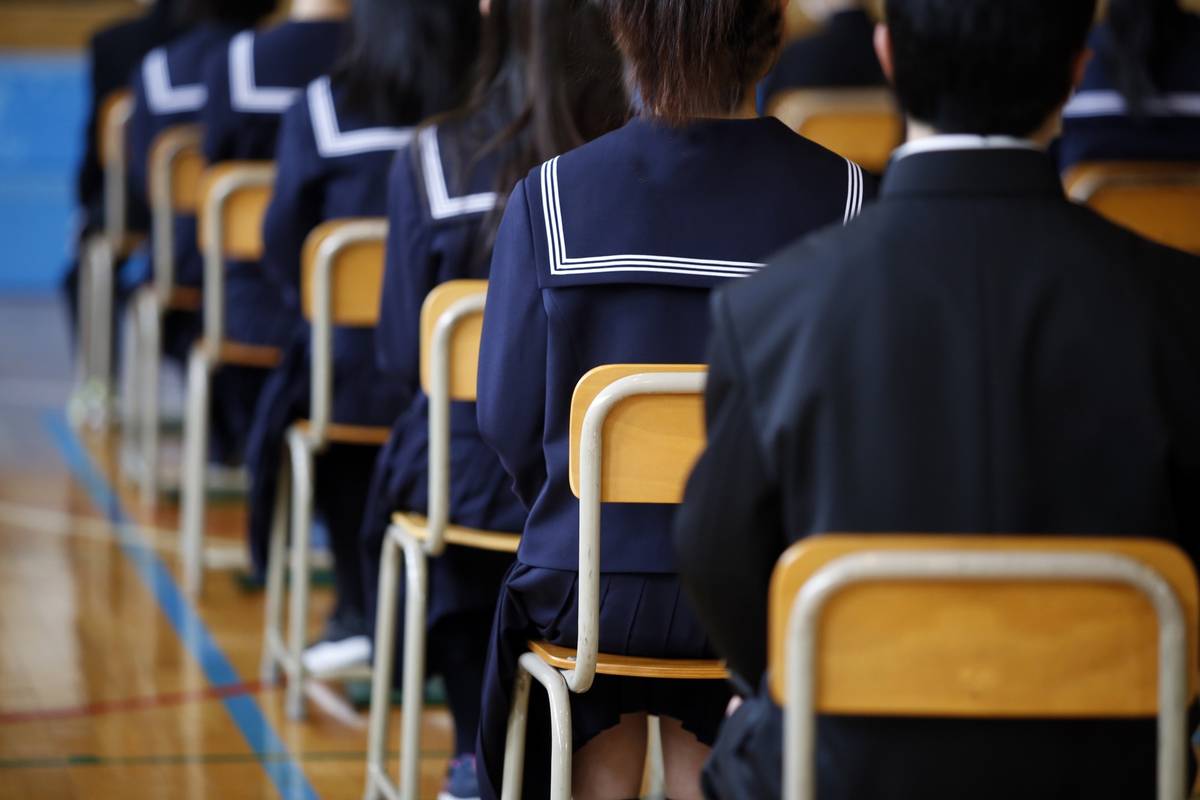 Students in school uniforms sitting in rows, viewed from behind, in a gym.