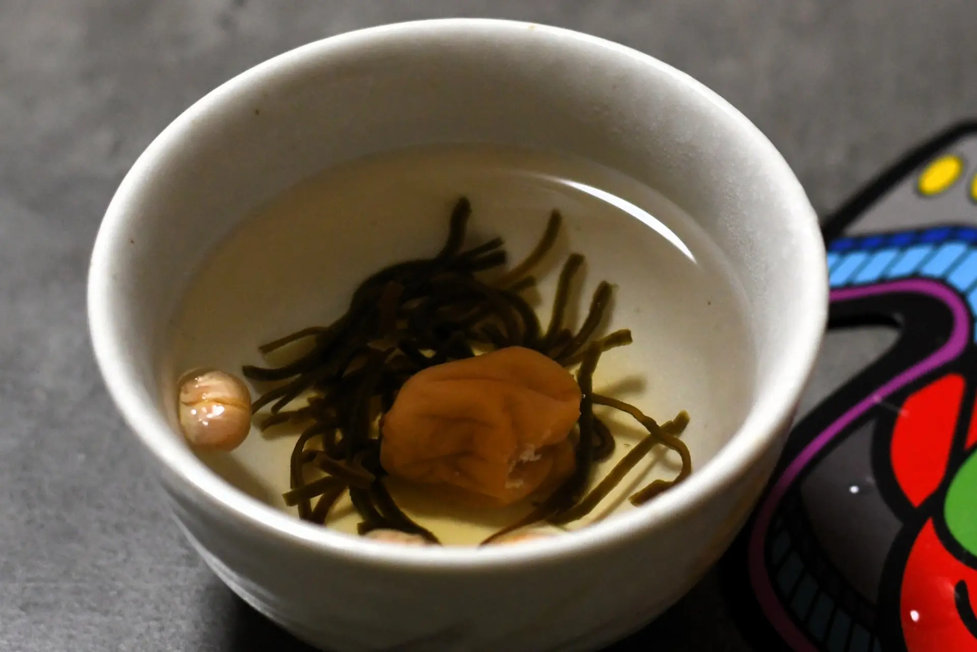 A white bowl containing a pickled plum, seaweed, and small beans in a clear liquid.